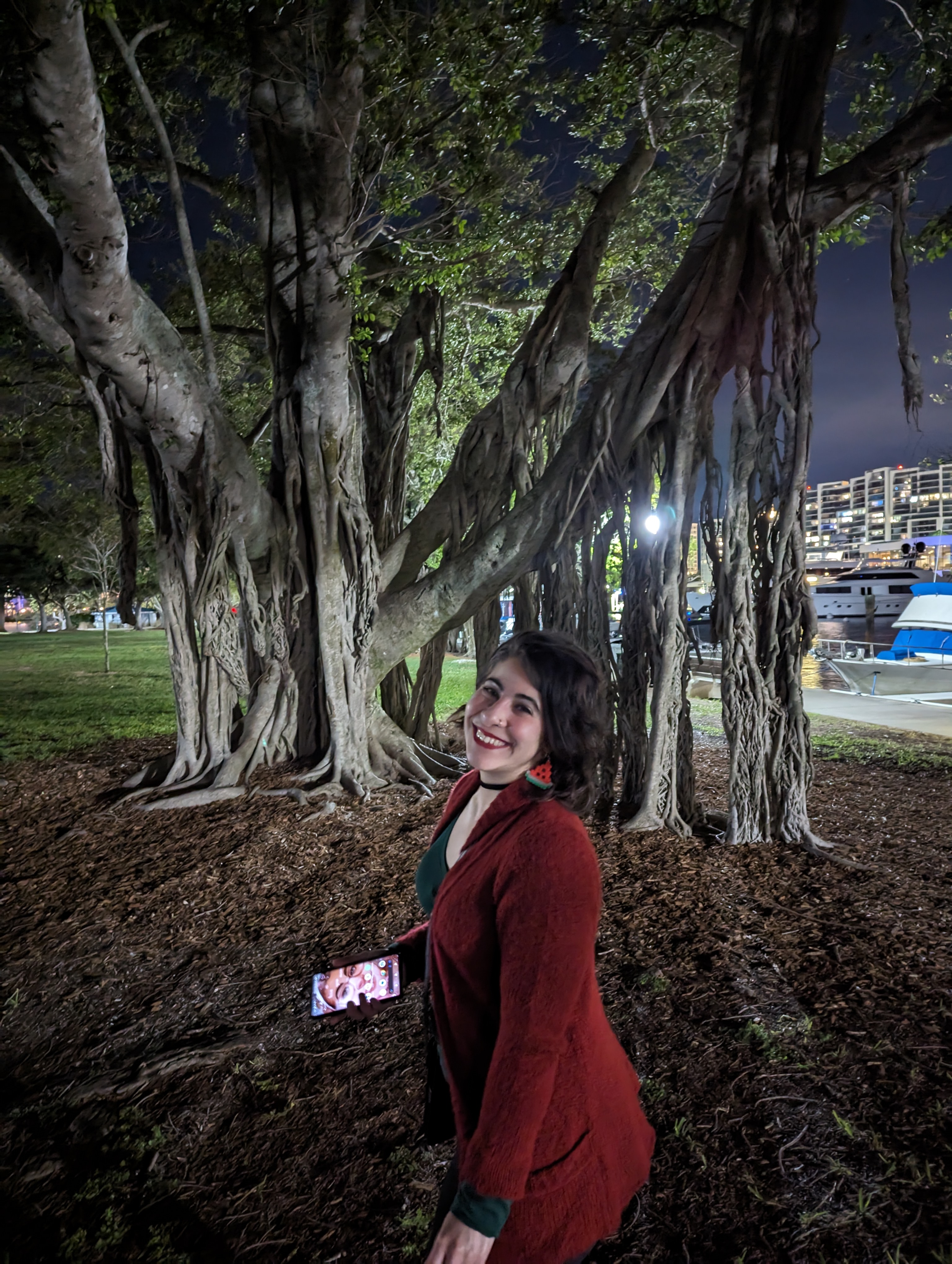 Photo of Eva Fiallos-Diaz in front of a banyan tree with ample roots. Eva is smiling and wearing a red jacket, green shirt, and watermelon earrings.